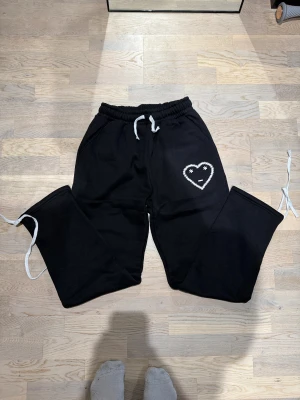 Carsicko mjukisbyxor  - Svarta mjukisbyxor från Carestick i 100% bomull, med vit snörning i midjan och ett broderat hjärta med ansikte på benet. Storlek M men passar L
