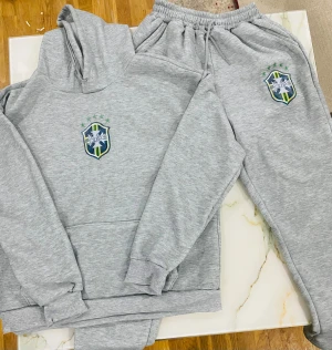 Grå hoodie med Saint-emblem - Grå hoodie med broderat Saint-emblem på bröstet, gröna stjärnor och detaljer i blått och gult. Tröjan har huva med snörning, stor magficka och ribbade muddar. Materialet är mjukt och känns som bomull eller polyester, perfekt för chill dagar.