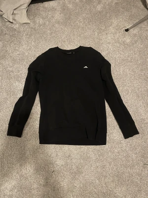 Svart sweatshirt från J.Lindeberg - Svart sweatshirt från J.Lindeberg med rund hals och diskret vitt logotryck på bröstet. Tröjan har långärmade ribbade muddar och är gjord i mjuk bomull. Enkel och stilren design som funkar till många olika looks.