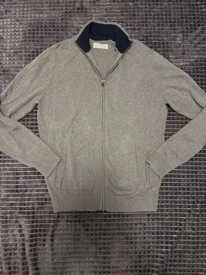 Grå kofta med dragkedja - Snygg grå kofta från Filmonte med hel dragkedja och ribbade muddar. Kragen har en mörkblå insida som ger en schysst kontrast. Perfekt för lager-på-lager och chill dagar. Två fickor framtill och mjukt material som känns skönt mot huden.