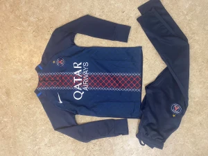 PSG långärmad fotbollströja Nike - Paris Saint-Germain långärmad fotbollströja från Nike i mörkblått med röda och vita detaljer. Tröjan har tryckt Qatar Airways-logga och PSG-emblem på bröstet. Materialet är lätt och andas, perfekt för träning. Säljes tillsammans med matchande byxor med PSG-logga.