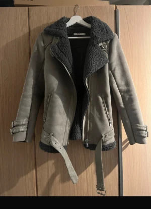 Grå teddyjacka med bälte och dragkedja - Säljer en grå teddyjacka från Gina Tricot med svart fluffigt foder, bred krage och coola detaljer som bälte i midjan och spännen vid ärmslut. Jackan har dragkedja framtill och två fickor med zip. Perfekt för dig som gillar streetstyle och vill hålla dig varm.☺️