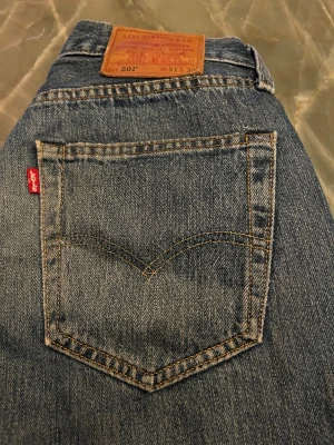 Levi's 501 blå jeans W31 L30 - Klassiska Levi's 501 jeans i blå denim med rak passform och normal midja. Snygga kontrastsömmar, fem fickor och ikonisk läderpatch baktill. Perfekta för dig som gillar tidlös stil och vill ha ett par jeans som alltid funkar. allting är bra skick, förutom det där lilla hålet