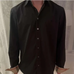 Svart  Burberry skjorta - Svart skjorta från Burberry med klassiska rutiga manschetter i beige, rött och svart. Den e perfekt för festliga stunder🍾🍾 kom privat för fler frågor🫡😀