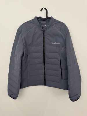 Lavandel Grå pufferjacka från Acne Studios storlek 46 (S) - Snygg grå pufferjacka från Acne Studios med stilren design och diskret logga på bröstet. Jackan har raka linjer, hög krage med knapp, hel dragkedja och quiltade paneler. Perfekt för dig som gillar minimalistisk stil och vill hålla dig varm. Vattentät.