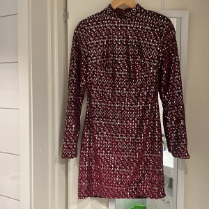Vinröd paljettklänning med öppen rygg - Perfekt för jul och nyår . Väldigt bra kvalitet på klänningen tjockt matrial. Vinröd kortklänning med långa ärmar, täckt av glittriga paljetter i ett randigt mönster. Klänningen har en hög hals och en djup, öppen rygg som ger en snygg kontrast. Materialet är syntet och passar perfekt för dig som vill sticka ut med en glammig look.