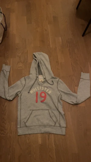 Grå hoodie Hollister med röd 19 - PRISET KAN DISKUTERAS. Bara att ställa frågor om det behövs. Snygg grå hoodie från Hollister i storlek M men känns som S med broderad text och siffran 19 i rött framtill. Tröjan har huva med snörning, magficka och långa ärmar. Perfekt för chill dagar och har en klassisk streetstil.