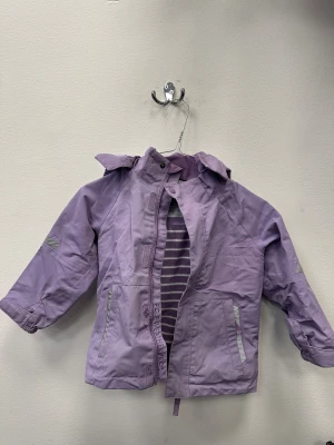 Ljuslila vindjacka från Polarn O. Pyret - Snygg ljuslila vindjacka från Polarn O. Pyret med avtagbar huva, reflexdetaljer på ärmar och fickor samt dragkedja och knappar framtill. Jackan har elastiska muddar vid ärmslut och är perfekt för blåsiga dagar. Insidan har ett randigt foder.