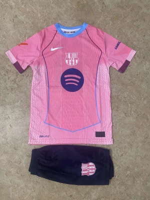 Barça Raphinha #11 rosa fotbollströja - Säljer en FC Barcelona fotbollströja med Raphinha och nummer 11 på ryggen. Tröjan är rosa med lila detaljer, korta ärmar och blå krage. Materialet är lätt och andas, Dri-Fit. Tryck med Spotify och UNHCR samt klubbmärke på både tröja och shorts.