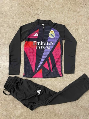 Real Madrid träningsset Adidas svart/lila - Säljer ett Real Madrid träningsset från Adidas med långärmad tröja och byxor. Tröjan har svart bas med grafiskt mönster i lila, rött och rosa samt klubbmärke och Adidas-logga. Byxorna är svarta med vit Adidas-detalj. Perfekt för fotbollsträning.