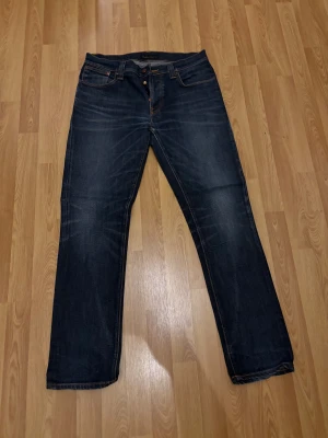 Mörkblå jeans från Nudie Jeans Co, 34/32 - Snygga mörkblå jeans från Nudie Jeans Co i klassisk straight fit. Jeansen har orange kontrastsömmar, fem fickor och knappgylf. Materialet är slitstarkt jeans och passformen är normal. Perfekt för dig som gillar tidlös stil och kvalitetsjeans.