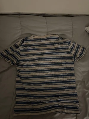 Randig t-shirt från Polo Ralph Lauren - Säljer en klassisk t-shirt från Polo Ralph Lauren i beige och blått med horisontella ränder. T-shirten har korta ärmar, rund halsringning och en bröstficka med broderad logga. Tillverkad i mjuk bomull för skön känsla.