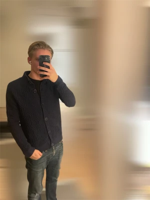 Mörkblå stickad cardigan med mönster - Säljer en mörkblå stickad kofta från Morris med snyggt diamantmönster och ribbade muddar. Koftan har hög krage och knäpps med stora knappar framtill. Perfekt för lager-på-lager