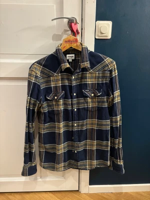 Rutig blå skjorta från Wrangler - Snygg rutig skjorta från Wrangler i storlek S. Skjortan är blå med beige och vita rutor, har klassisk krage, två bröstfickor med knappar och långa ärmar. Perfekt för dig som gillar avslappnad stil med lite western-vibe.