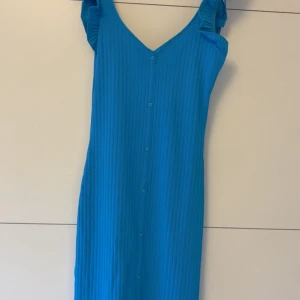 Blå ribbad långklänning med volang - Snygg blå långklänning med ribbad struktur och knappar framtill. Klänningen har v-ringad hals och axelband med volangdetaljer som ger en cool touch. Materialet är mjukt och stretchigt, perfekt för sommarens varma dagar.