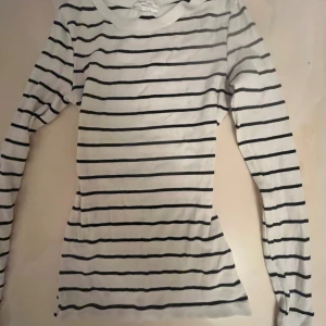 Randig långärmad tröja från Ginatricot - Vit långärmad tröja från Ginatricot med svarta horisontella ränder. Tröjan är ribbad och har en klassisk, smal passform som sitter snyggt på kroppen. Perfekt för en enkel och stilren look.