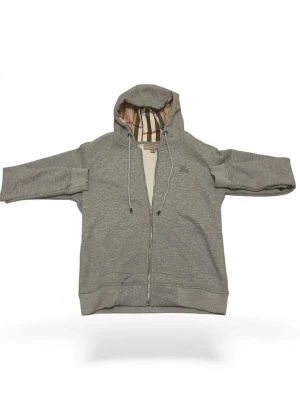 Grå hoodie från Burberry med dragkedja - Snygg grå hoodie från Burberry med klassiskt rutigt foder i huvan och broderad logga på bröstet. Tröjan har hel dragkedja, snörning i huvan och ribbade muddar. Perfekt för dig som gillar stilrena och exklusiva plagg.