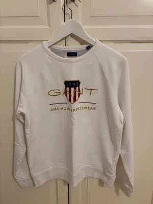 Vit sweatshirt från GANT - Vit sweatshirt från GANT med broderad logga och emblem i guld, rött och blått på bröstet. Tröjan har rund hals, ribbade muddar och lång ärm. Perfekt för en sportig och clean stil.
