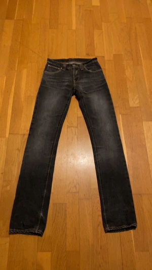 Svarta Nudie Jeans Super Slim  - Säljer ett par svarta jeans från Nudie Jeans, modell Super Slim. De har klassisk femficksdesign, gul söm på bakfickorna och är tillverkade i 100% bomull. Passformen är slim och benen är raka med en mörk tvätt. De är lite tajta i midjan men det är ingenting som märks. 