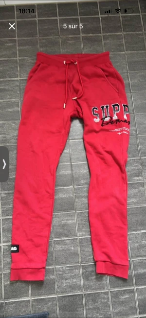 Röda mjukisbyxor Supply Demand - Röda mjukisbyxor med resår och snörning i midjan. Byxorna har tryck med texten 'SUPER' och detaljer på ena benet samt ribbade muddar vid bensluten. Perfekta för chill eller träning.