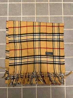 Rutig halsduk från Burberrys i ull - Klassisk rutig halsduk från Burberrys i 100% ull. Halsduken har fransar i ändarna och den ikoniska Burberrys-etiketten. Perfekt accessoar för att lyfta din outfit. Anledningen att de stör ”burberrys” är för att de är en äldre modell.