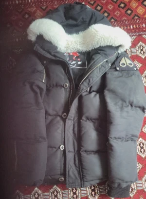 Svart pufferjacka Moose Knuckles - Svart pufferjacka från Moose Knuckles med ikonisk logga på ärmen och broderad hockeypatch på insidan. Jackan har en stor huva med fluffig vit fuskpäls, dragkedja och knappar framtill samt ribbade muddar vid ärmslut. Perfekt för kalla dagar.