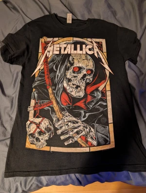 Svart Metallica t-shirt med dödskalle - Svart t-shirt med stor Metallica-print framtill. Trycket visar en dödskalle med röda ögon och en lie, samt en mindre dödskalle med röd snörning. T-shirten har rund hals och korta ärmar, tillverkad i mjuk bomull. Perfekt för dig som gillar Metal och Metallica, och är i bra skick!