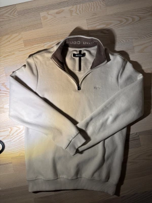 Beige quarter zip from Hugo Boss - Snygg beige långärmad tröja från Hugo Boss med halv dragkedja och broderad logga på bröstet. Kragen har en brun insida med broderad Hugo Boss-text. Mjuk bomullskvalitet och ribbade muddar för extra komfort. Perfekt för chill dagar.