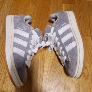 Adidas Campus grå sneakers 41 1/3 - Adidas Campus sneakers i grå mocka med klassiska vita ränder på sidorna och vit logga på plösen. Skorna har vit snörning och en ljusbrun gummisula med mönster. Stilren och tidlös modell som passar perfekt till jeans eller shorts.