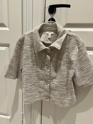 Beige croppad skjorta med guldknappar - Snygg croppad skjorta från H&M i beige och vitt med ett rutigt, strukturerat tyg. Skjortan har korta ärmar, klassisk krage och fyra guldiga knappar framtill som ger en lyxig touch. Aldrig använd 