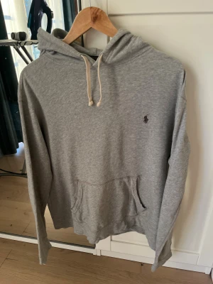 Grå hoodie från Polo Ralph Lauren - Grå hoodie från Polo Ralph Lauren med klassisk logga på bröstet. Tröjan har huva med snörning, magficka och långa ärmar. Tillverkad i mjuk bomullsmix som är skön att ha på sig. Perfekt för chill dagar och enkel att matcha med jeans eller joggers. Mycket bra skick förutom ett litet hål på armbågen men som inte märks så mycket