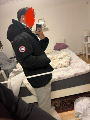 Svart dunjacka från Canada Goose - Svart dunjacka från Canada Goose med ikonisk röd och vit patch på ärmen. Jackan har huva, knappar framtill och två stora fickor. Perfekt för kalla dagar och har en clean, klassisk look som funkar till allt. Snygg passform och riktigt varm.