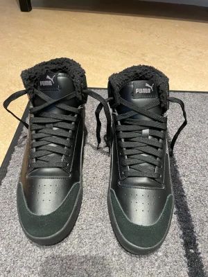 Svarta höga sneakers från Puma - Säljer ett par svarta höga sneakers från Puma med snyggt syntetläder och mockadetaljer. Skorna har snörning, rund tå och en fluffig, pälsliknande foderkant som gör dem extra sköna och varma. Perfekt för dig som gillar streetstyle och vill ha något som sticker ut. Perfekt för vinter andrig använt dom 