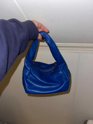 Blå axelväska med dragkedja - blå axelväska i mjukt skinnimitation med silverfärgad dragkedja och justerbar axelrem. 