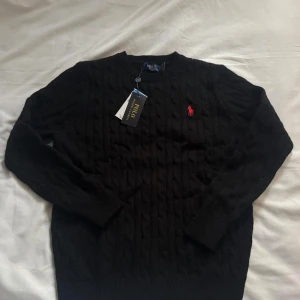 Svart kabelstickad tröja Polo Ralph Lauren - Svart kabelstickad tröja från Polo Ralph Lauren med klassisk rund hals och röd broderad logga på bröstet. Tröjan har långa ärmar och ribbade muddar vid ärmslut och nederkant. Perfekt för dig som gillar stilren och tidlös design.