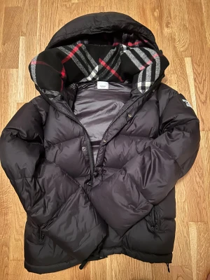 Svart Burberry pufferjacka convertible med luva - Svart pufferjacka från Burberry med stor huva och klassiskt rutigt foder i svart, vitt och rött. Jackan har dragkedja och knappar framtill, samt Burberry London England-logga på ärmen. Fylld med dun och har en loose passform, perfekt för kalla dagar.