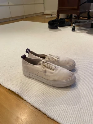 Vita eytys sneakers med lila detaljer - Säljer ett par vita sneakers med platt sula och rund tå. Skorna har snörning med guldfärgade öljetter och en lila hälflik som snygg kontrast. Tillverkade i bomullstyg, perfekta för dig som gillar enkel och clean stil.