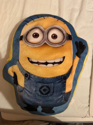 Minionkudde - Bilderna visar en Minion-mjukdjur/kudde, formad som en karaktär från filmen Despicable Me. Produkten är färgglad och ser ut att vara i gott skick utan synligt slitage. Perfekt som prydnad eller mysig kudde för barn och fans av Minions.