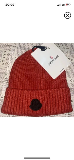 Röd ribbad mössa från Moncler - Snygg röd ribbstickad mössa från Moncler med svart logopatch framtill. Mössan har uppvikt kant och är tillverkad i mjukt stickat material som håller dig varm och ger en cool vibe.