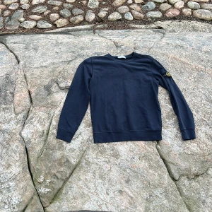 Mörkblå Stone Island sweatshirt - Snygg mörkblå sweatshirt från Stone Island med klassisk rund hals och ikonisk logga på vänster ärm. Tröjan är i mjuk bomull och har långa ärmar med ribbade muddar. Perfekt för dig som gillar streetwear och vill ha en clean look.