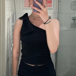 Svart glittrig oneshoulder topp med rosett - Säljer en tjusig svart glittrig oneshoulder topp med stor rosett på axeln. Toppen är croppad och i storlek 146/152 från Kappahl. 