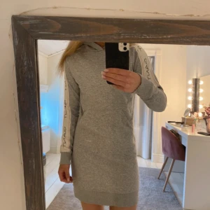 Grå hoodieklänning från Gant - Superskön grå hoodieklänning från Gant med långa ärmar och huva. Klänningen har vita ränder med Gant-logga längs ärmarna och en stor ficka framtill. Perfekt för chill dagar och har en sportig vibe.