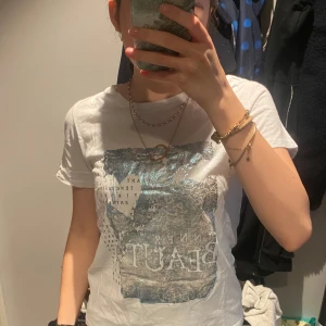 Vit t-shirt med metallictryck - Vit t-shirt med korta ärmar och ett stort, glansigt metallictryck i silver och blått framtill. Trycket har text och grafiska detaljer som ger en cool och modern vibe. Klassisk rund halsringning och mjukt material som känns skönt mot huden.