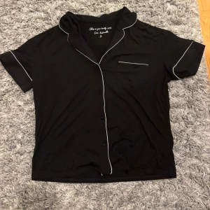 Svart kortärmad skjorta med vita detaljer - Svart pyjamas skjorta med kort ärm och vita kontrastsömmar längs med kragen, ärmslut och ficka. Klassisk krage, bröstficka och knäppning framtill. Skön och luftig pyjamas. 