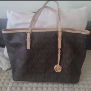 Brun Michael Kors axelväska med logomönster - Stor brun axelväska från Michael Kors med klassiskt MK-logomönster över hela väskan. Väskan har ljusbeiga handtag i skinn och en snygg guldfärgad MK-detalj hängande framtill. Perfekt för dig som vill ha plats för allt och ändå se stilren ut.