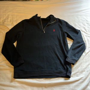 Svart half zip tröja Polo Ralph Lauren - Svart långärmad tröja från Polo Ralph Lauren med klassisk röd broderad logga på bröstet. Tröjan har en snygg half zip-dragkedja och ribbade muddar. Perfekt för dig som gillar stilren och enkel design.
