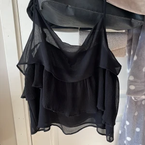 Svart plisserad linnetopp - Säljer en svart linnetopp från Monki i storlek Large men skulle säga att den är liten i storleken så passar även medium. Toppen har tunna axelband med knyt, plisserade volanger och är tillverkad i ett lätt, transparent material som ger en luftig känsla. Perfekt för dig som gillar lager på lager och coola detaljer, värt att tillägga att den inte är så genomskinlig som den ser ut på bilden🖤