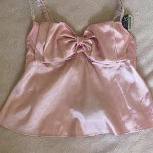 Rosa satinlinne med rosett 🎀 - Säljer ett glansigt rosa satinlinne med en söt rosett framtill, linnet har justerbara axelband 💕