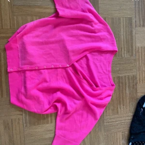 Knallrosa kofta med knappar - Säljer en knallrosa kofta med rund hals och knappar framtill. Koftan har långa ärmar och är gjord i ett mjukt, tunt material som känns skönt mot huden. Perfekt för dig som vill ha en färgstark och enkel tröja att slänga på dig.