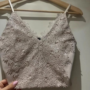 Beige spetslinne med smala axelband - Supersnyggt beige linne med tunna axelband och v-ringning. Toppen är täckt av detaljerad spets med blommigt mönster och har en croppad passform. Perfekt för dig som vill ha en feminin och trendig look.